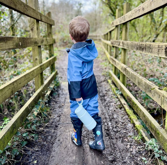 Child wearing blue waterproofs and holding a non-toxic stainless steel waterproof bottle whilst crossing a bridge during a nature walk