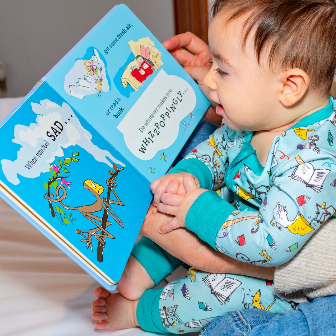 Image of a baby wearing a Ducky Zebra's Roald Dahl 'The Giraffe and the Pelly and Me' Sleepsuit reading Roald Dahl's 'Fantastic Feelings' book