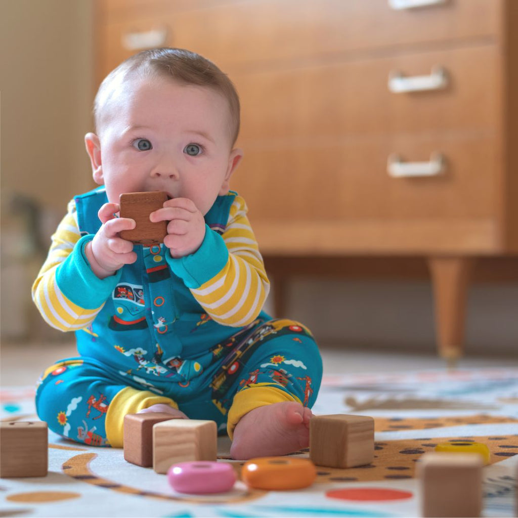 Baby sitting up wearing Ducky Zebra teal dungarees and a yellow and white stripe bodysuit below. The baby is holding a block in its hands 
