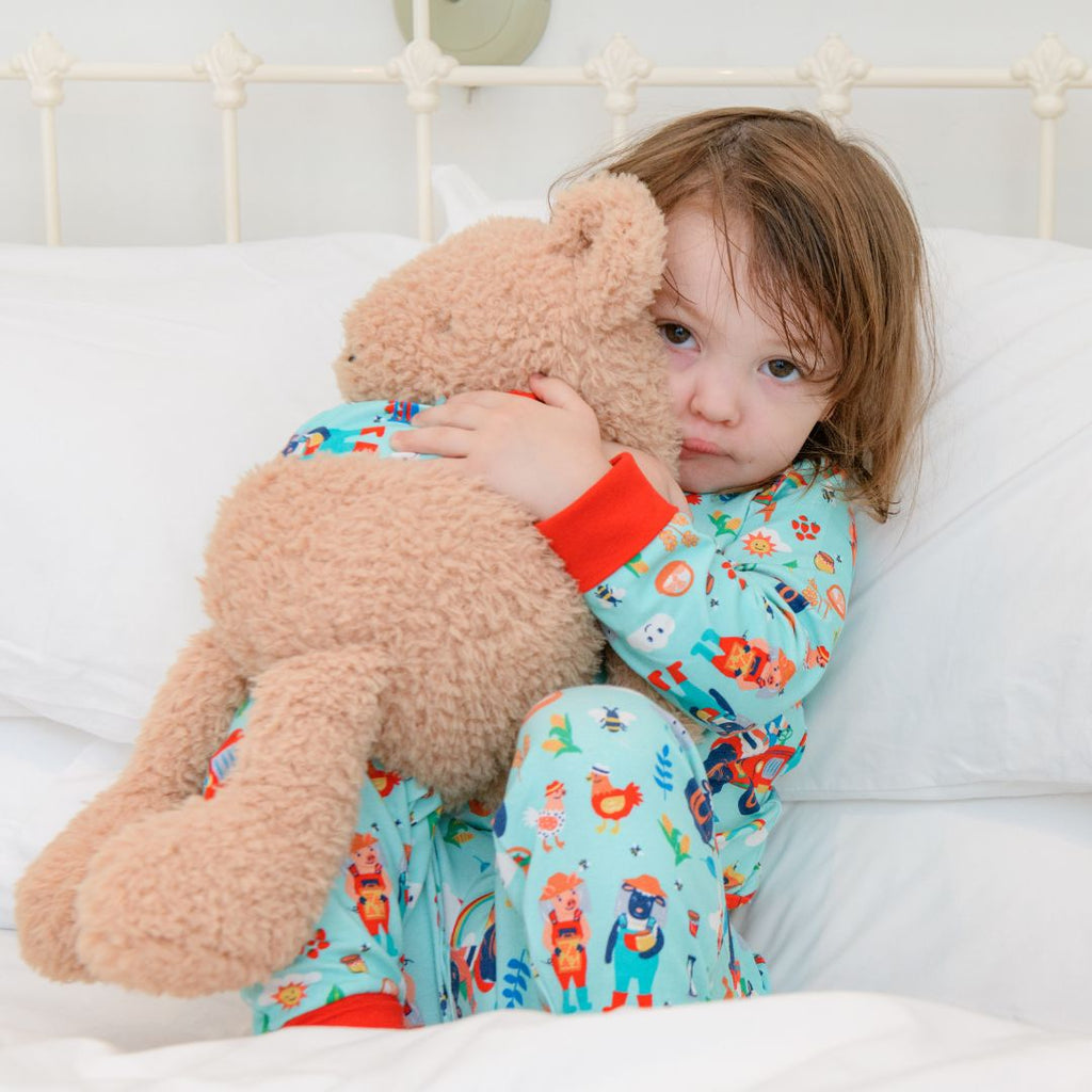Image of a girl snuggling a teddy in bed wearing Ducky Zebra sustainable pyjamas with a fun repeat print of a pig and sheep on the farmyard. 