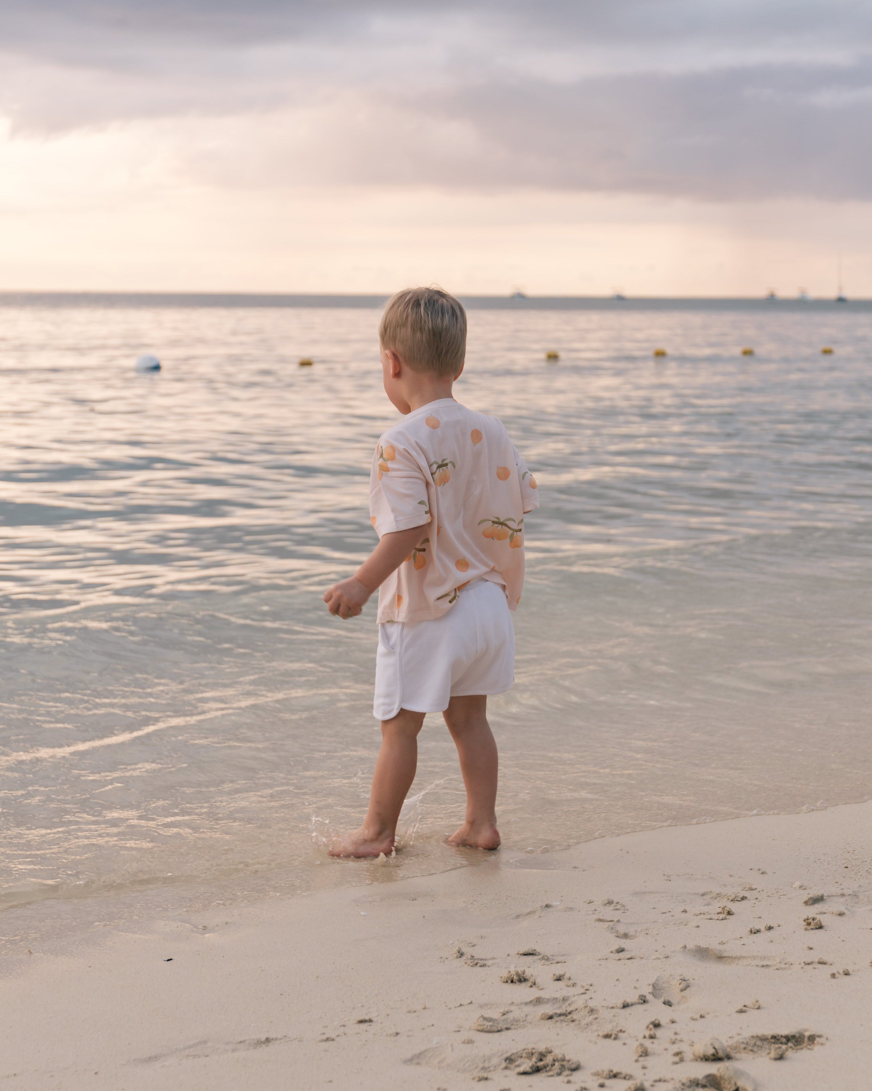 Toddler on the beach at sunset wearing Wildgreen’s Apricot T-shirt from the Island Life collection. Made from 100% GOTS-certified organic cotton, this gender-neutral kids tee features a soft apricot-hued print, perfect for eco-conscious families seeking sustainable and stylish clothing. #OrganicKidswear #SustainableFashion