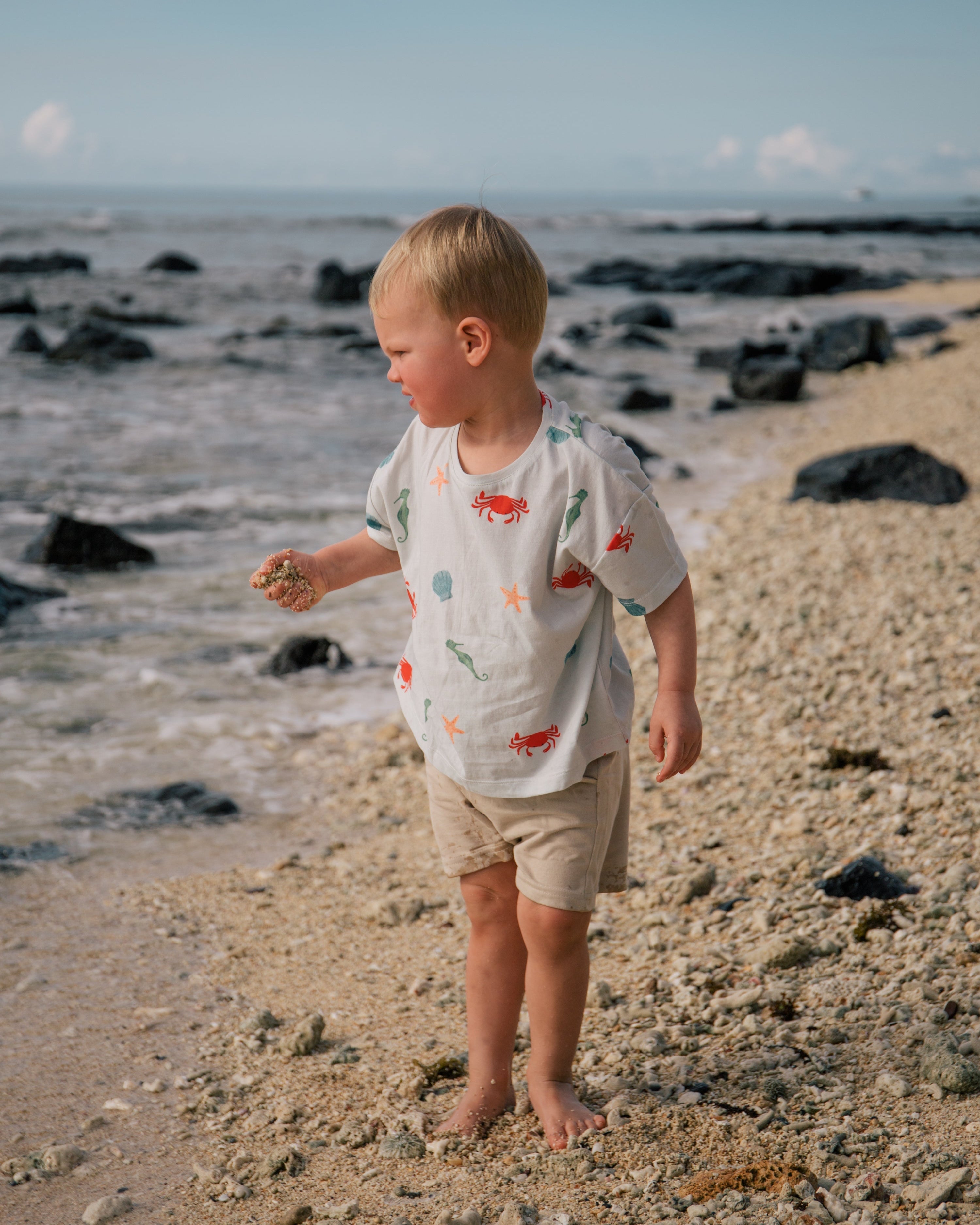 Toddler on a tropical beach wearing an organic Marine T-shirt from Wildgreen’s Island Life collection. Ethically made from 100% GOTS-certified organic cotton, this gender-neutral kids tee features a vibrant ocean-inspired print. Perfect for eco-conscious families seeking stylish and comfortable clothing