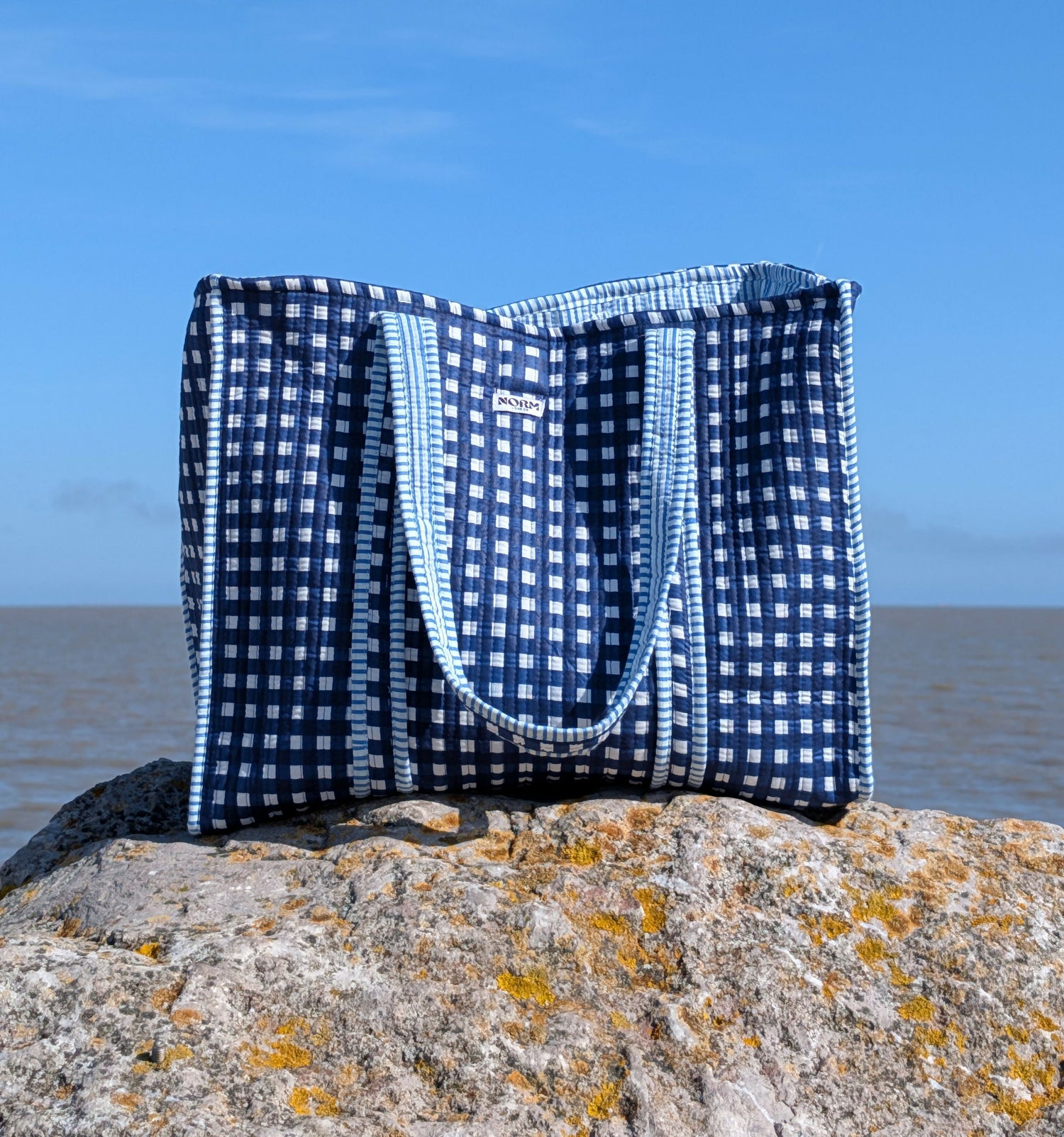 blue chequered tote bag with handles sitting on a rock on the beach with the sea and the sky as the backdrop
