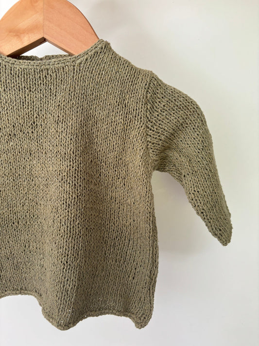 A green, knit silk sweater for babies displayed on a wooden hanger.