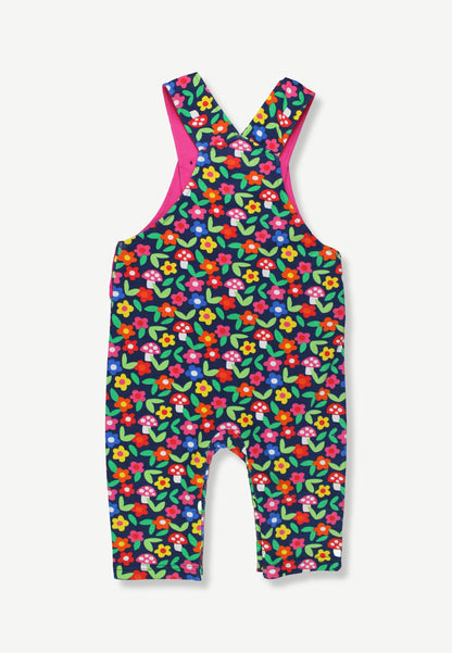 A baby romper with an all-over colorful floral print on a dark green background, featuring bright pink shoulder straps and a matching pink inner lining, shown against a light gray background.