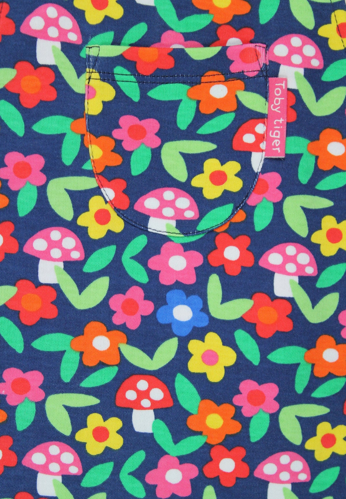 A close-up of a fabric product featuring a colorful floral and mushroom print on a navy blue background. A curved pocket is stitched into the upper left area, with a small pink fabric brand tag reading 'joy tufei' attached near the pocket edge.