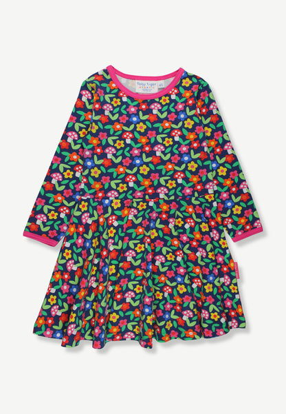 A long-sleeve tiered dress for toddlers or young girls, featuring a dense multicolor floral print on a dark green background with bright pink, red, yellow, and blue flowers, finished with a contrasting pink collar and cuffs.