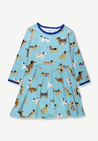 A light blue long-sleeve dress for toddlers, featuring an all-over playful print of various dog illustrations. The dress has a pleated skirt and is finished with contrasting dark blue trim at the crew neck and cuffs.