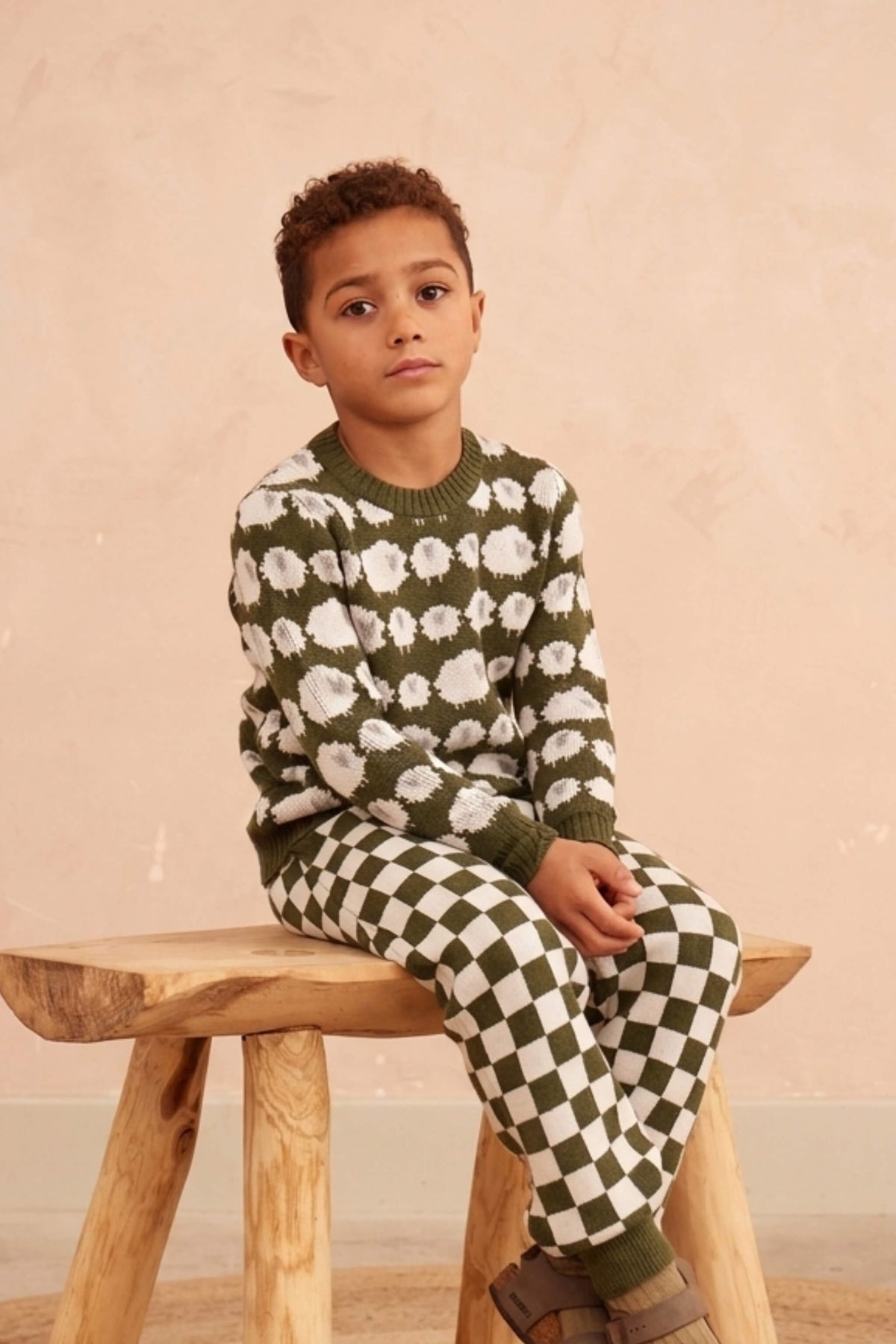 Boy standing against a plaster wall in the Flock of Sheep Jumper in avocado green