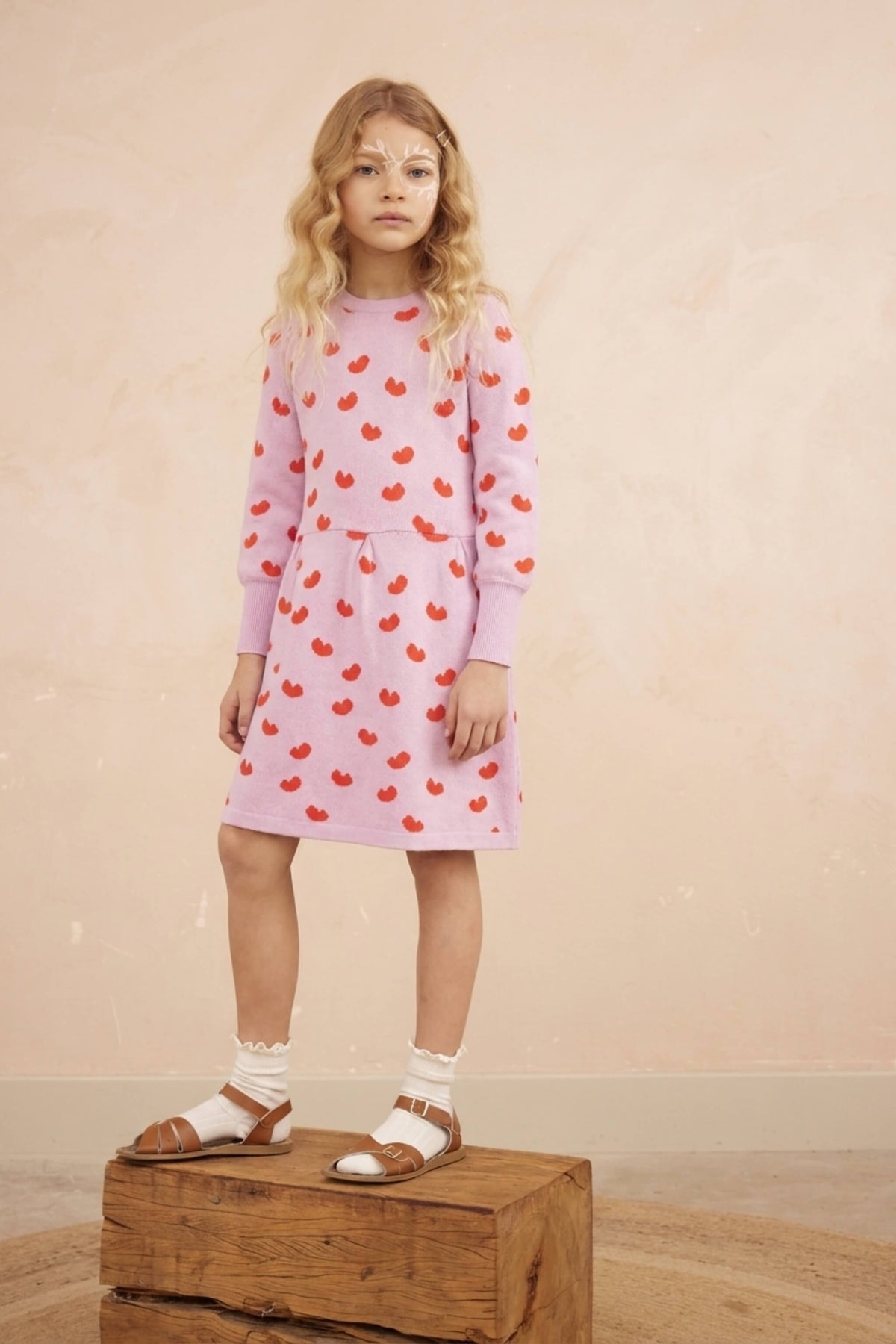 Girl standing against a plaster wall in the Love Hearts Dress in bubble gum pink