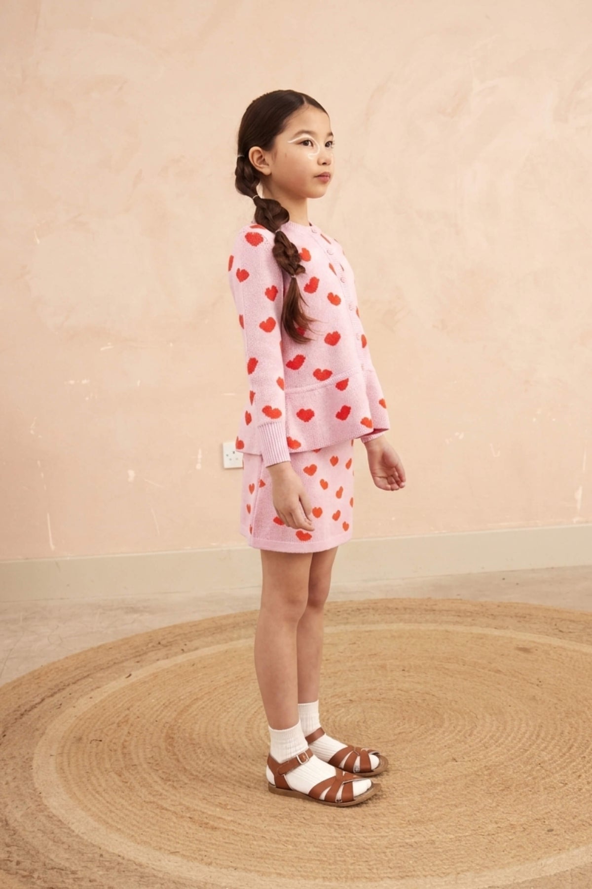 Girl in the Love Hearts Peplum Cardigan in bubble gum pink against a plaster wall