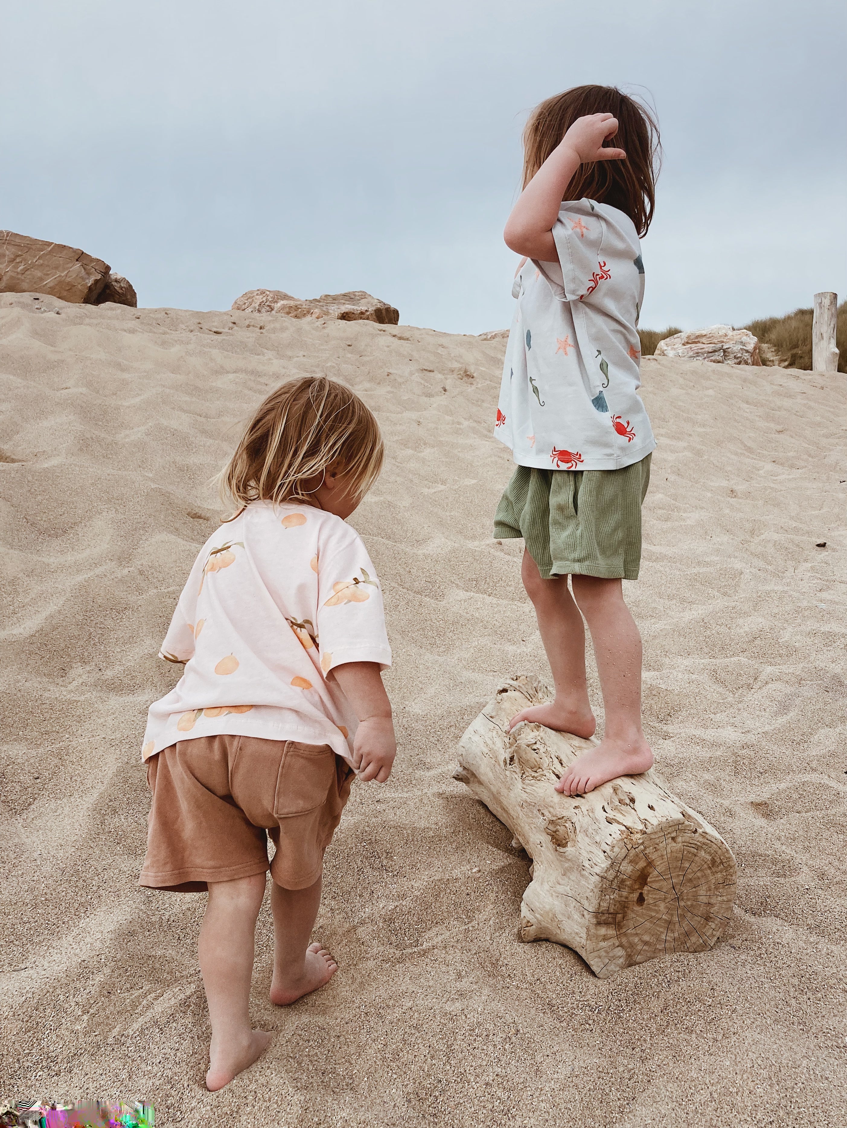 Children playing on a beach wearing Wildgreen organic cotton, gender-neutral t-shirts designed for sustainable, outdoor play. Ocean print and apricot peach pattern.