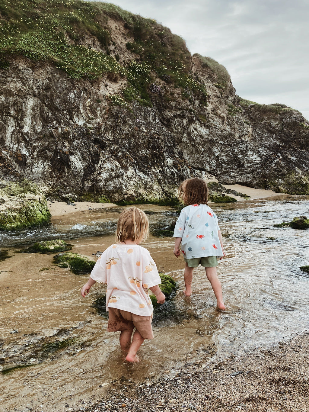 Two children playing in a beach river wearing Wildgreen organic cotton, gender-neutral t-shirts designed for sustainable, outdoor play.